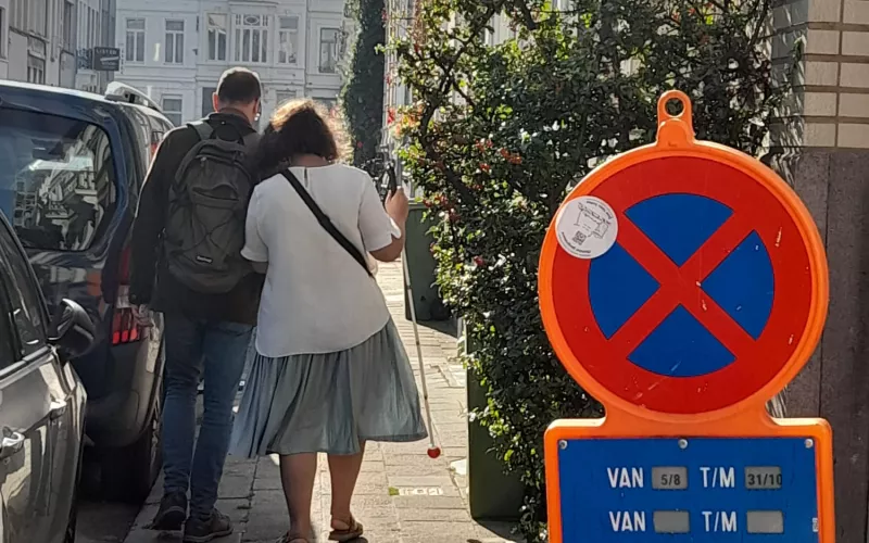 foto met vooraan een parkeerbord in de weg van de stoep en een sticker erop, op de achtergrond een vrouw met witte stok begeleid door een man