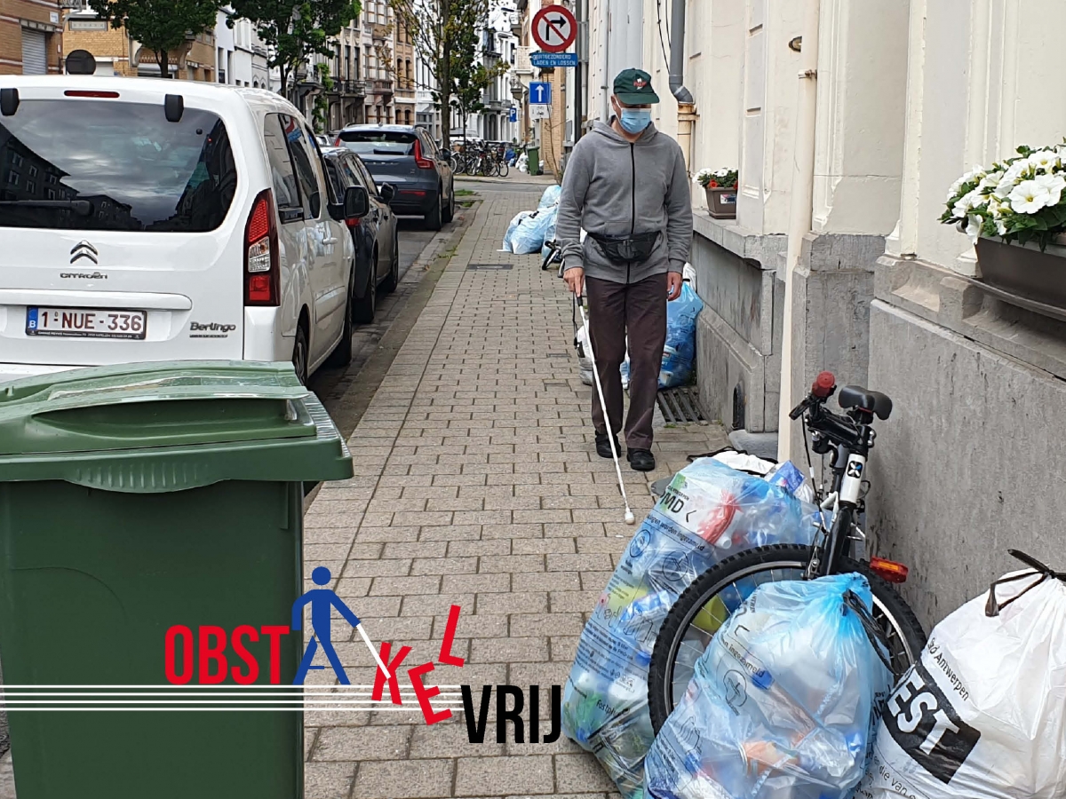 Foto van Bart op de stoep met zijn witte stok en allerlei obstakels (vuilnis, fiets, vuilnisbak)