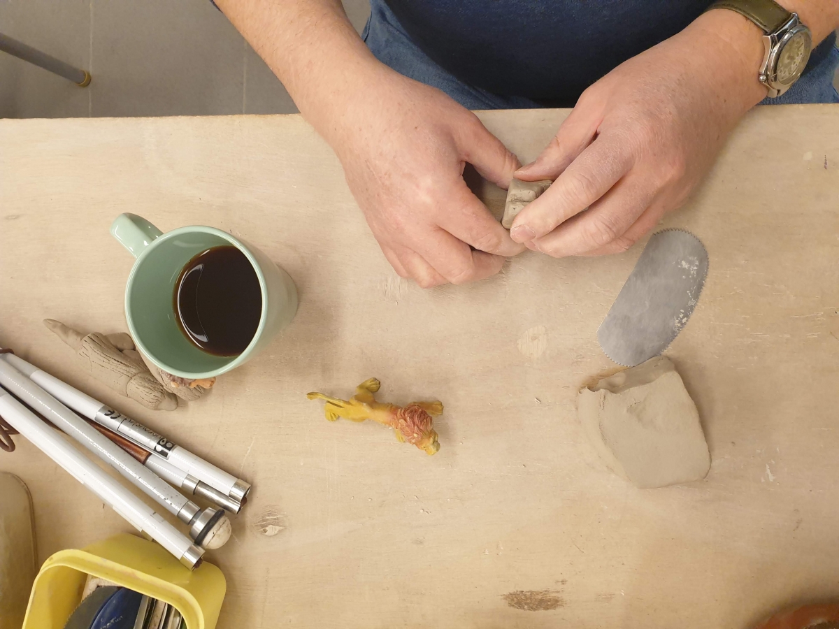 Een tafel in het atelier met 2 handen in de klei, een tas koffie en een witte stok